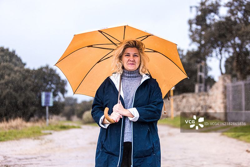 穿着蓝色和灰色羊毛高领毛衣的女人手持黄色雨伞遮挡雨水，正看向镜头。水，冬天，秋天，淋湿和下雨。图片素材