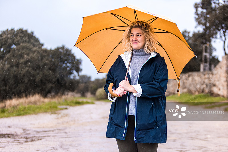 穿着蓝色和灰色羊毛高领毛衣的女人手持黄色雨伞遮挡雨水，侧身望去。水，冬天，秋天，淋湿和下雨。图片素材