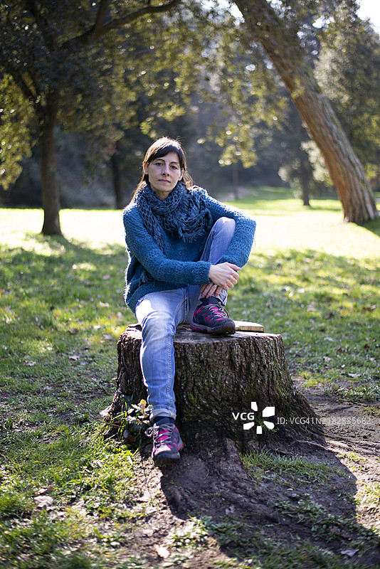 一位西班牙裔女性坐在砍伐的树干上，放松地看着镜头，身处大自然中。图片素材