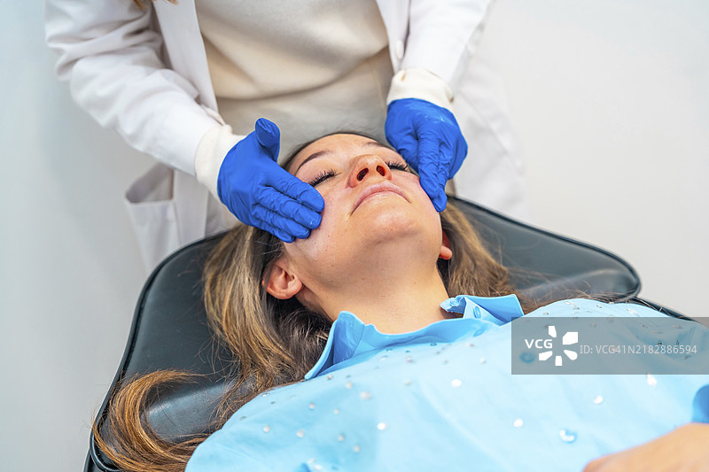 美丽的女医生在诊所为一名女性的脸部涂抹麻醉膏，以便在美容治疗中注射透明质酸。图片素材