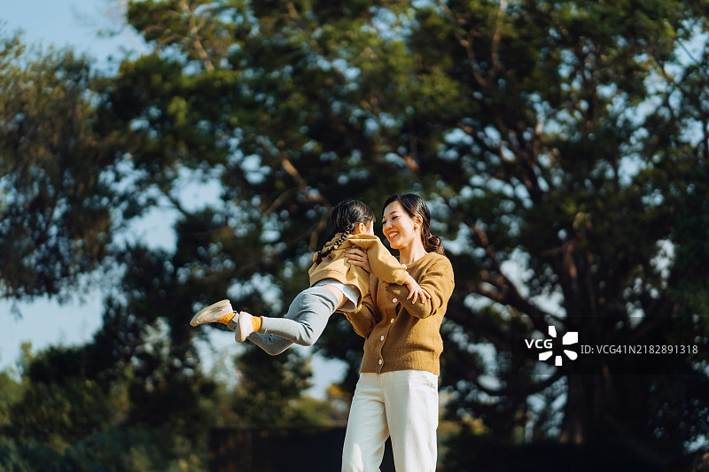 无忧无虑的年轻亚洲母亲在公园里转动可爱的小女儿，享受户外时光，享受大自然的乐趣。健康的家庭生活方式，爱与亲密的概念。图片素材