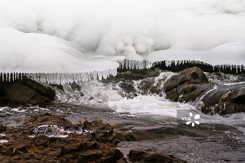 韩国京畿道的河流中，快速流动的水面上覆盖着雪和冰，冰柱像牙齿一样悬挂在上方，河床岩石。图片素材