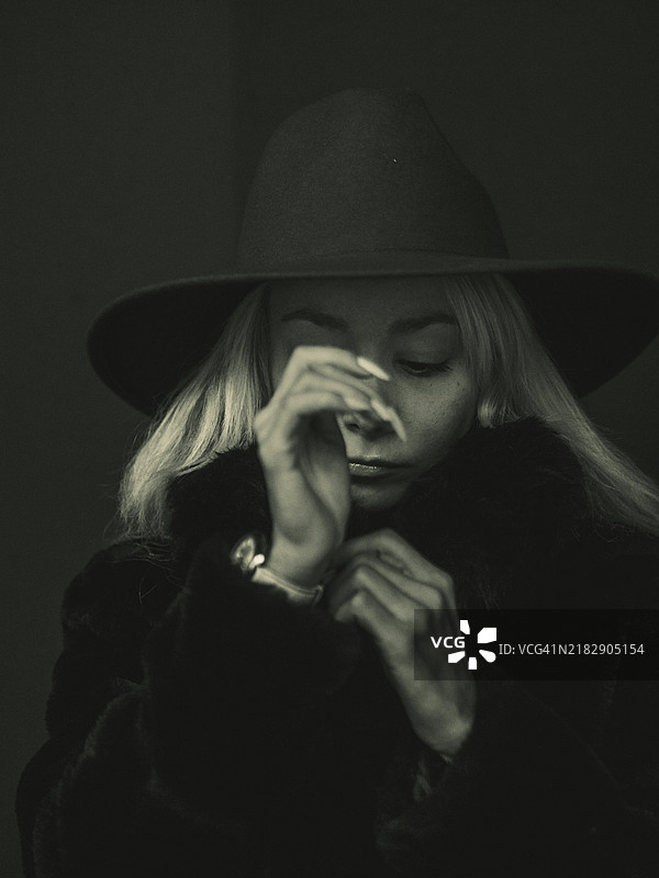 一幅单色图像，描绘了一位沉思的金发女性，戴着帽子，脸部部分被手遮挡。图片素材