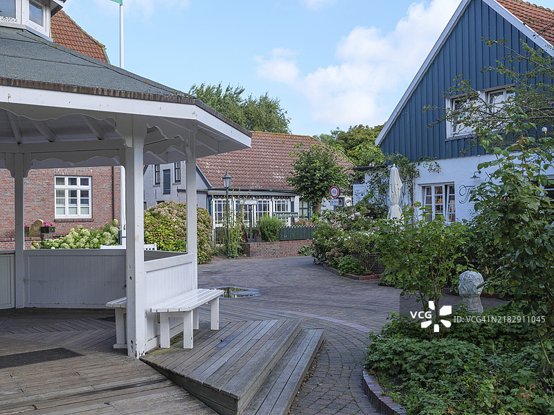 阳光明媚的日子里，德国东弗里西亚斯皮克罗克的村庄街道，街道旁有一个凉亭和几座被绿色花园环绕的房屋。图片素材