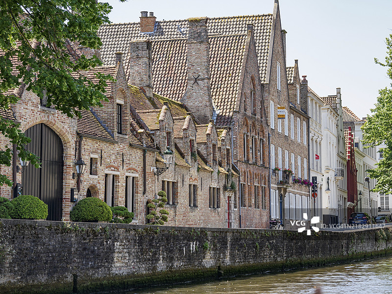 比利时布鲁日,欧洲,夏日晴朗天气下运河旁的历史砖房和修剪整齐的灌木丛,古老的历史房屋外立面,带有塔楼和桥梁。图片素材