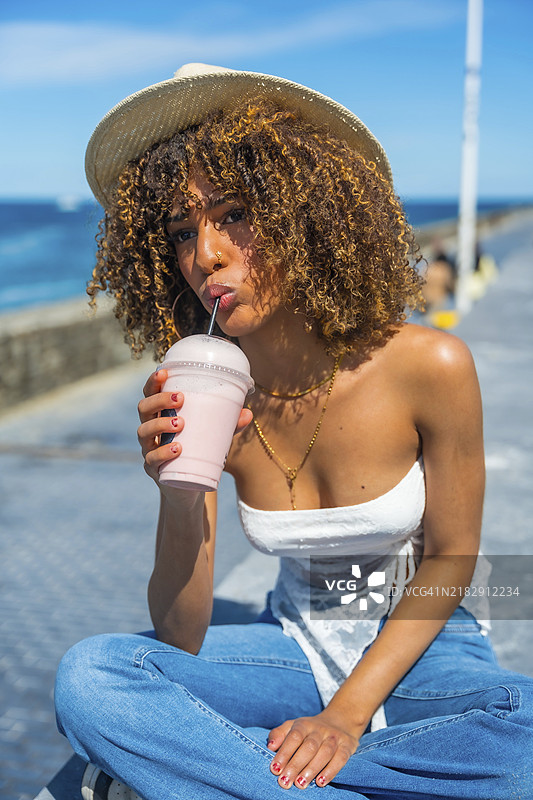 一位拉丁裔年轻酷炫且性感的女性坐在海边，喝着奶昔的竖版照片图片素材