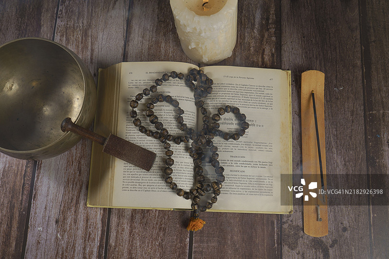 从上方俯视一本《博伽梵歌》书籍，旁边有念珠、藏式唱碗、蜡烛和香在木桌上。图片素材