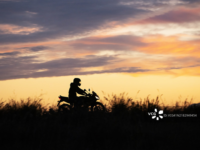 日落时分，天空下骑摩托车的剪影男子在田野上。图片素材