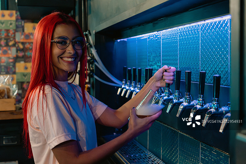 微笑的红发酒保在现代酒吧的蓝色灯光下从酒龙头倒啤酒图片素材