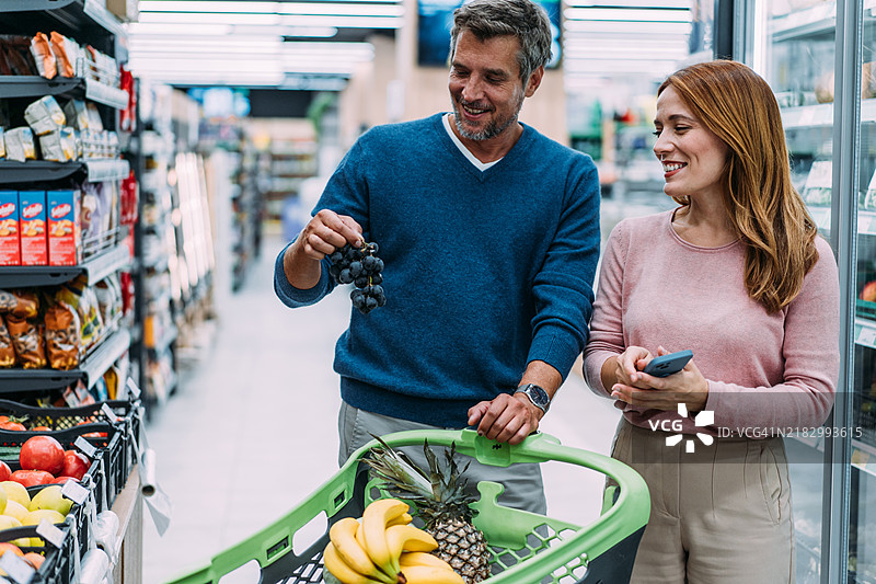 微笑的情侣在超市里推着购物车挑选商品。图片素材