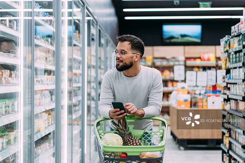 快乐的男人在超市购物时选择商品并使用手机。图片素材