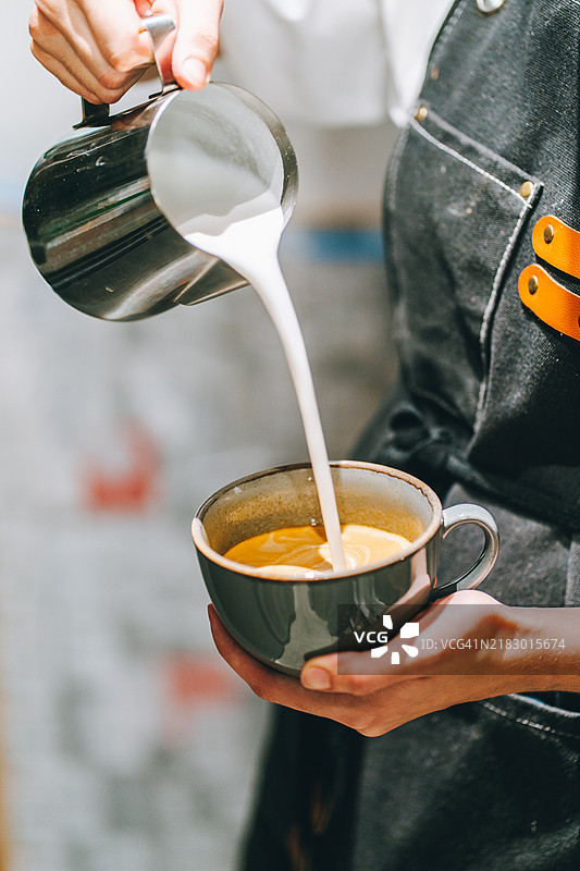 穿着围裙的咖啡师在浓缩咖啡中倒入热牛奶，制作泡沫丰富的卡布奇诺。图片素材
