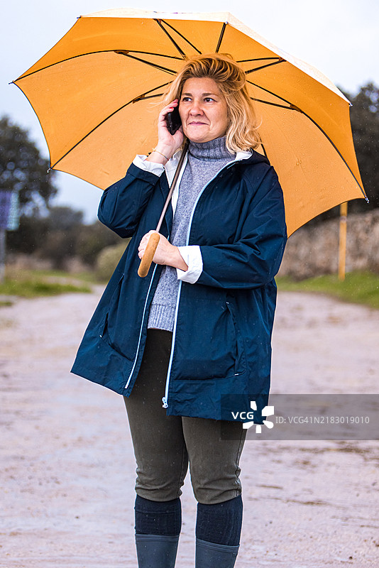 一位穿着雨衣、高领毛衣和雨靴的女性在乡村风景中，手持黄色雨伞遮挡雨水，同时在用手机通话，画面为中景。图片素材