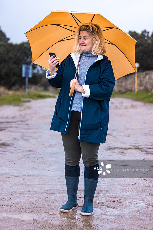 在雨天的乡村风景中，一位穿着雨衣、高领毛衣和雨靴的女性，手持一把黄色雨伞以保护自己免受雨淋，同时查看她的手机。图片素材