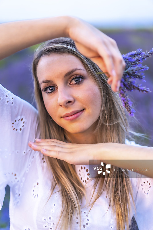 一位年轻的金发白人女性穿着白色连衣裙，站在西班牙纳瓦拉的精心培育的薰衣草田中。生活方式，乡村生活。手中拿着一束紫色花朵，享受乐趣。图片素材