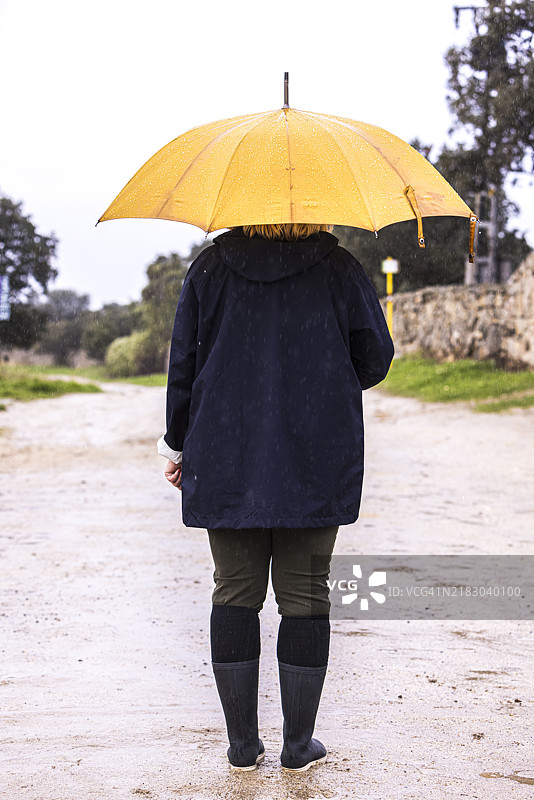 一位穿着蓝色雨衣、手持黄色雨伞的女性背影远景，遮挡着雨水。水，冬季，秋季，淋湿和下雨。图片素材