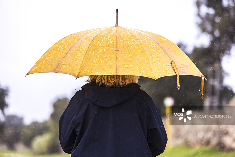 一位穿着蓝色雨衣、手持黄色雨伞的女性背影特写，遮挡着雨水。水，冬季，秋季，淋湿和下雨。图片素材