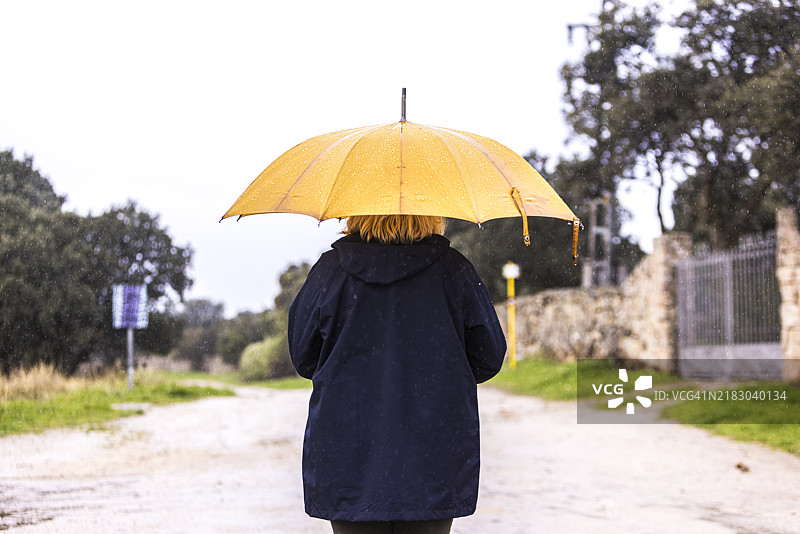 一位穿着蓝色雨衣、手持黄色雨伞的女性背影特写，遮挡着雨水。水，冬季，秋季，淋湿和下雨。图片素材
