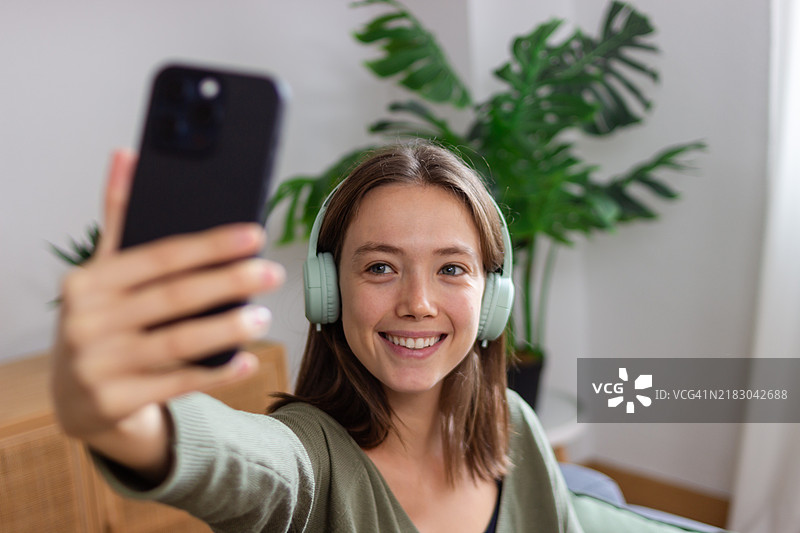年轻女性在家中戴着耳机用智能手机自拍图片素材