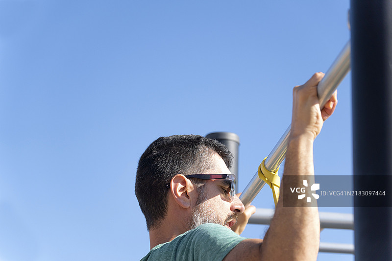 戴着太阳镜的胡子男人从背后看，在户外健身房用弹性健身带做引体向上训练背部。图片素材