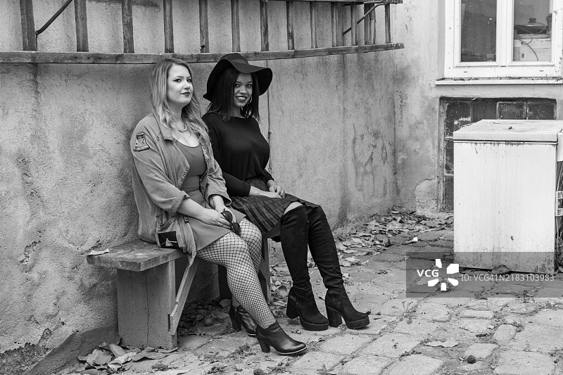 黑白照片中，两位女性朋友坐在后院的长椅上。一位年轻女性肤色较白，长着金色长发，另一位是非裔美国人，戴着一顶非常大的帽子。周围环境显得有些荒废。图片素材