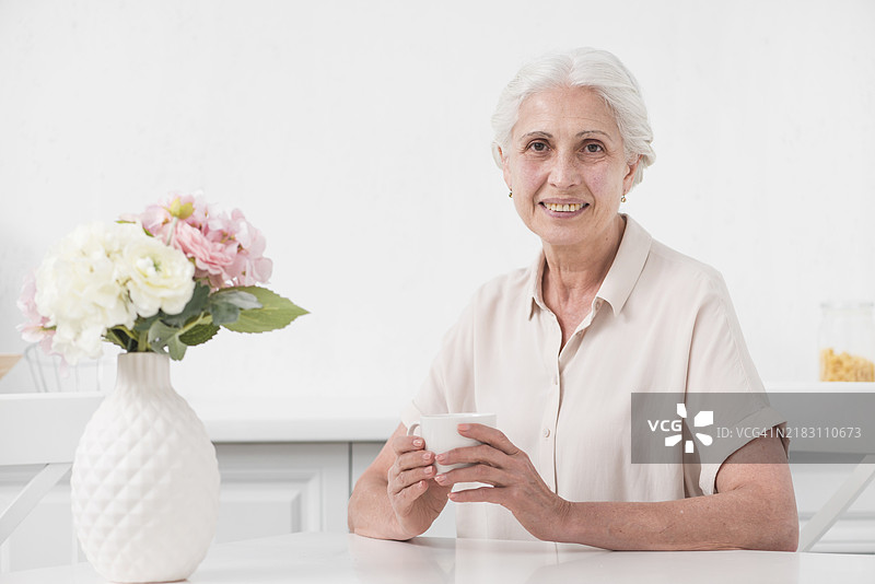 一位年长女性手持咖啡杯，桌上有花瓶和白色桌布。图片素材