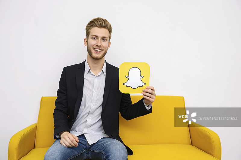 年轻男子坐在黄色沙发上，手持Snapchat图标图片素材