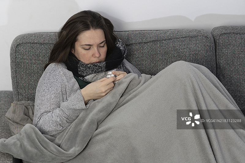 生病的女人在家里的沙发上盖着毯子，服用退烧药。图片素材