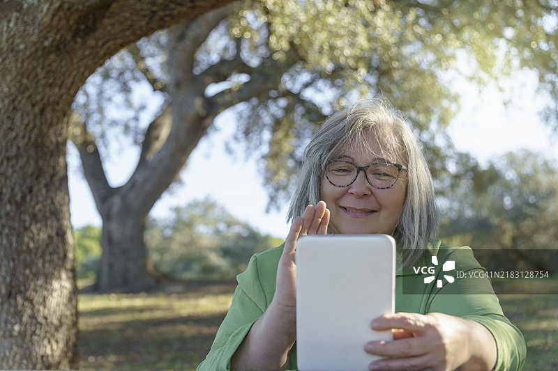 一位成熟的白发女性戴着眼镜，在阳光明媚的乡村里通过数字平板电脑进行视频通话，正挥手致意。图片素材