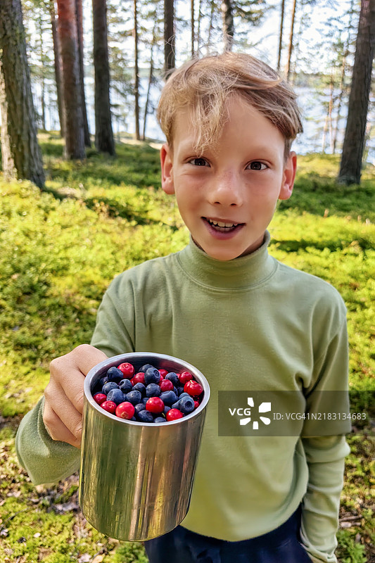 微笑的男孩穿着绿色卫衣，手里拿着一个装满新鲜蓝莓和红越橘的金属杯，站在阳光明媚的湖边森林空地上。图片素材