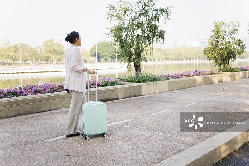 亚洲女商人在公园里推着手提行李箱走。图片素材