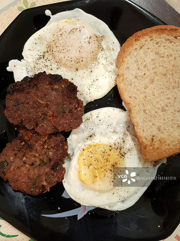 平铺视角的煎蛋、羊肉饼（羊肉提基）和一片面包，摆放在桌子上的盘子里，适合健康早餐。图片素材