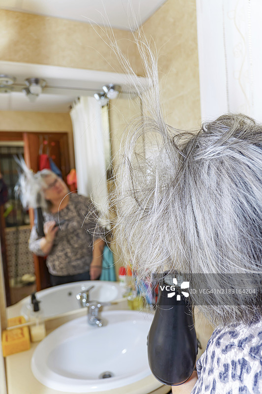 成熟的白发女性在镜子前用手持吹风机吹干头发图片素材