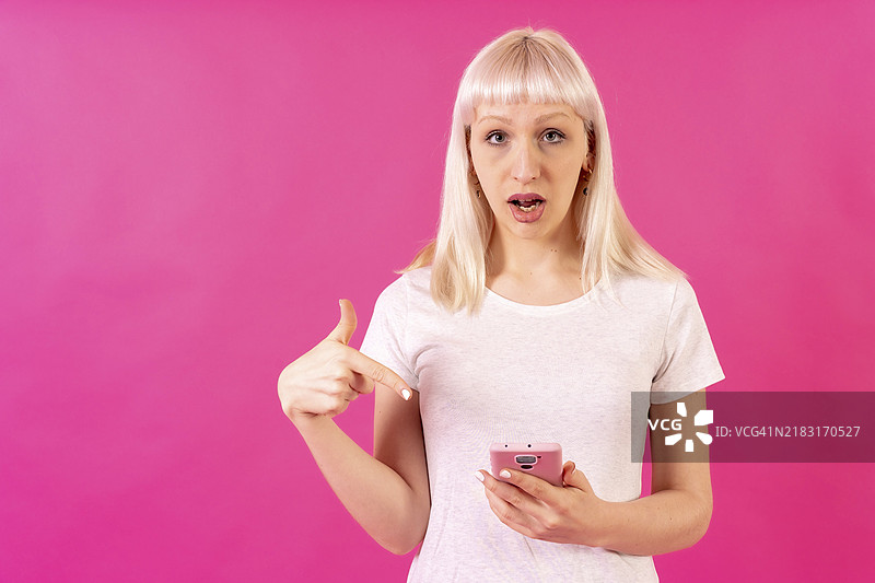 金发白人女孩在粉色摄影棚背景前，指着手机，留有空白空间。图片素材