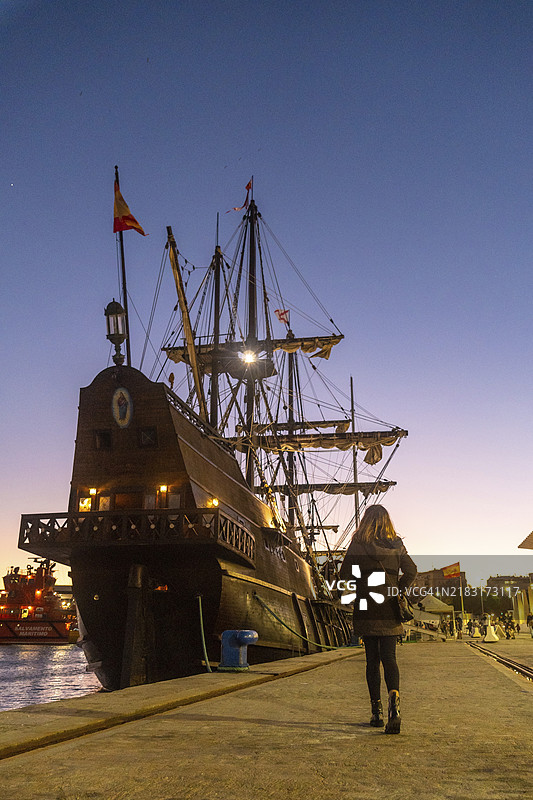 一位游客在西班牙安达卢西亚马拉加市的马拉加港Muelle Uno的海滨长廊上，日落时分参观老船。图片素材