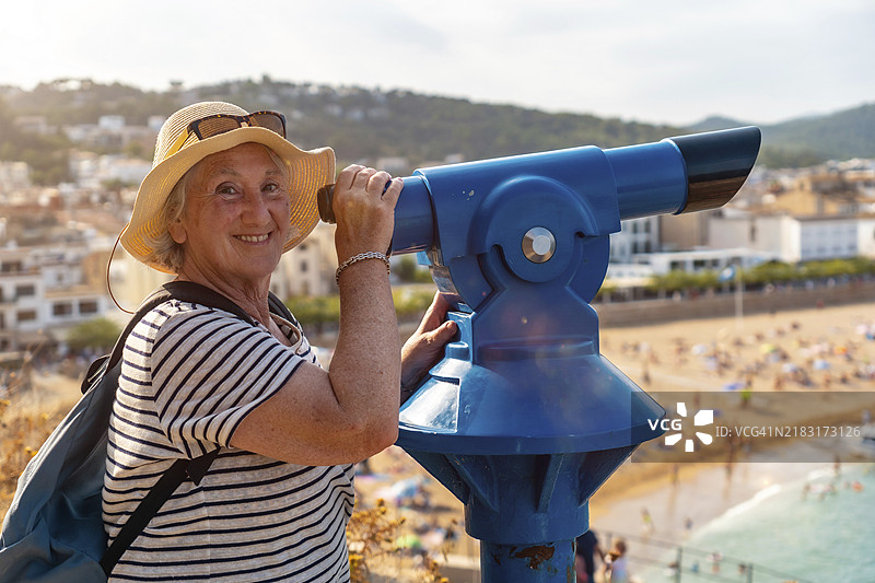 侧面视角的快乐老年女性正在通过投币望远镜观看并微笑图片素材