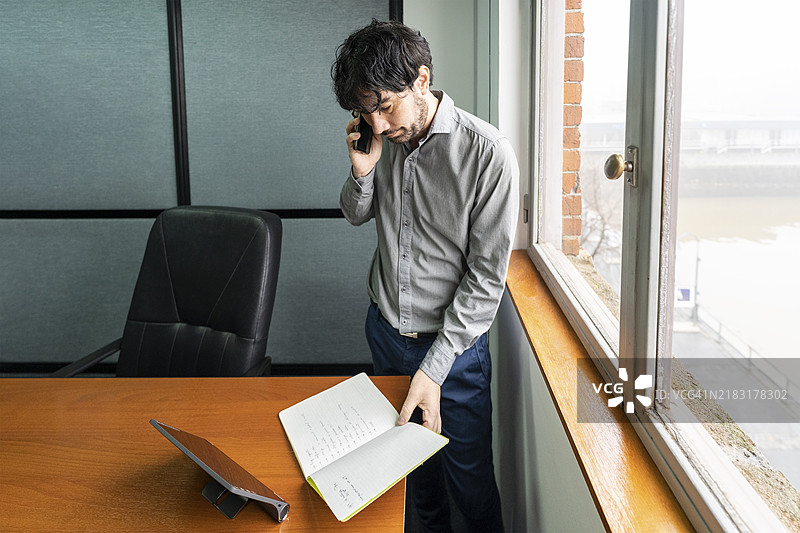 在办公室工作的拉丁裔商人，俯瞰着河流。他正在打电话并查看笔记。图片素材