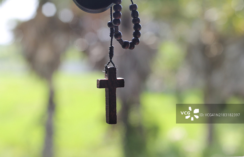 一串木制念珠，十字架悬挂在模糊的自然背景前。图片素材