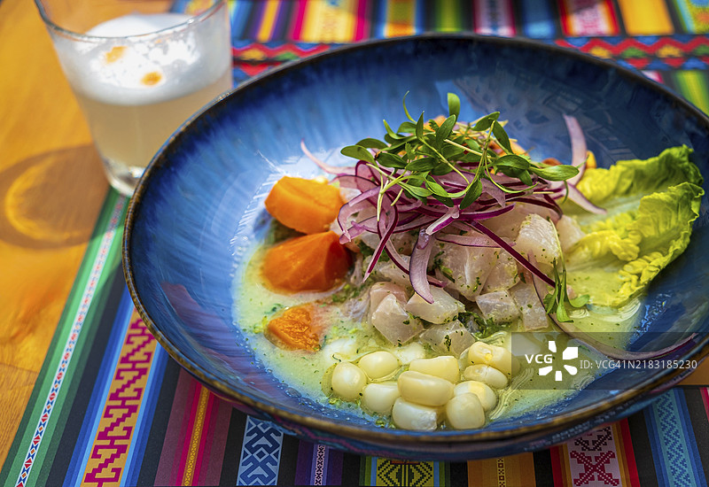 新鲜的生鱼片用蓝色盘子盛放,放在带有传统图案的餐垫上,旁边是一杯皮斯科酸鸡尾酒。选择性对焦,背景模糊。秘鲁风味的生鱼片由生鱼、红洋葱、玉米、烤玉米和红薯制成。图片素材