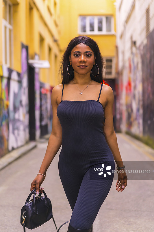 时尚的城市风格，黑肤女孩穿着黑色西装和高筒靴，在城市街道上漫步，手提黑色包包。图片素材