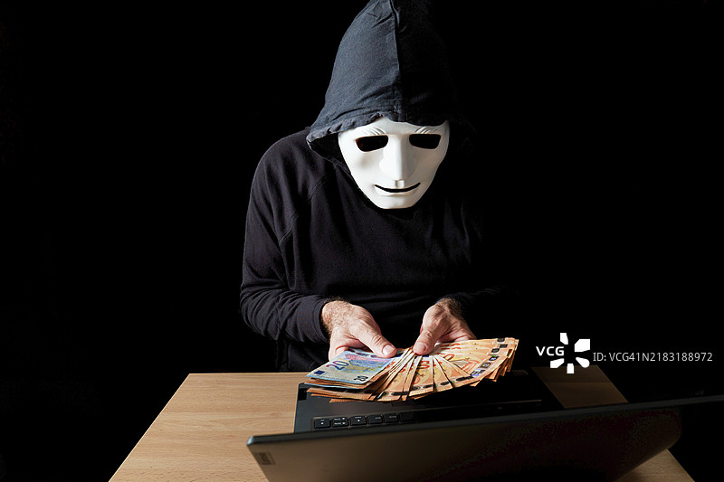 身穿黑色衣服、戴着白色面具和黑色兜帽的黑客正在手中数着欧元钞票，网络犯罪概念，背景为黑色。图片素材