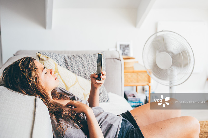 在炎热的夏天，悲伤的女人在家中用手机和电风扇。图片素材
