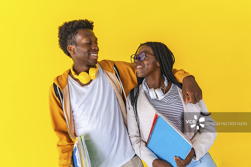 两名非洲大学生朋友在黄色背景下的工作室肖像，男生和女生相互拥抱，微笑着站在一起。图片素材
