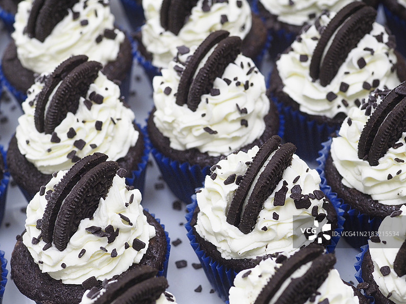 一组不健康的黑巧克力杯子蛋糕，上面装饰着甜美的奶油霜和饼干，放在蓝色纸杯里。图片素材