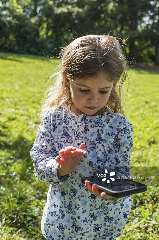 一个小女孩手持智能手机，静思地站在阳光明媚的草地上图片素材