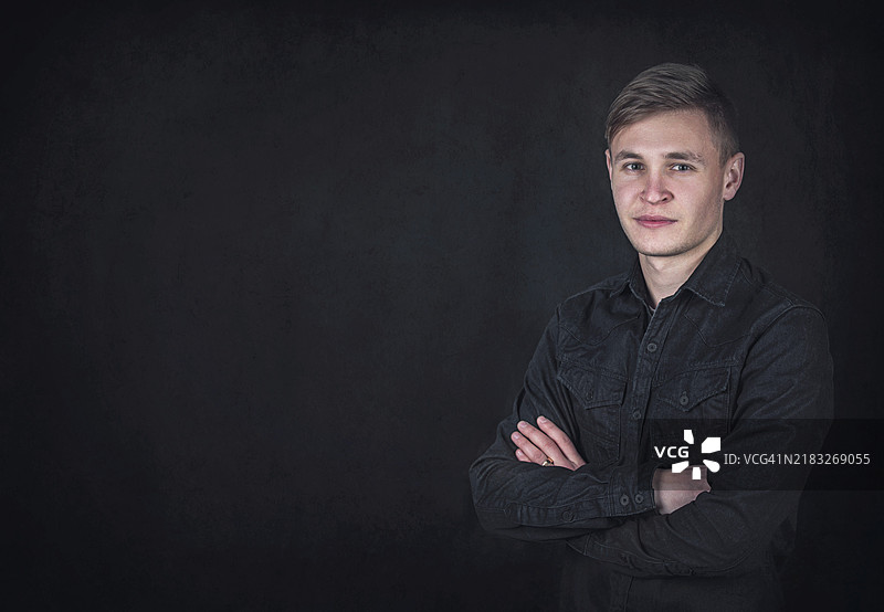 自信年轻男子的肖像，双臂交叉，背景为黑色墙壁，留有空白空间。成功的男性学生，面部表情坚定，穿着深蓝色牛仔夹克。图片素材