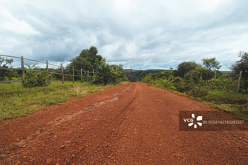 一条红土路延伸向乡村，四周环绕着树木、绿意盎然的植物和木栅栏，天空阴云密布。柬埔寨蒙多基里，亚洲图片素材