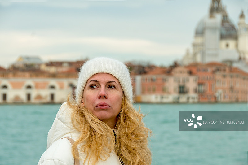 一位五十多岁的金发女性，头戴毛线帽，面露担忧神情，特写于威尼斯。图片素材