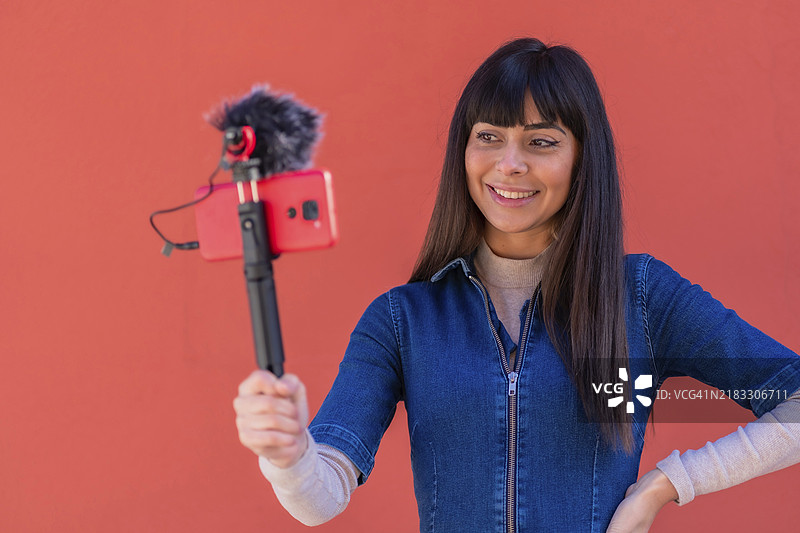 棕发女孩穿着蓝色牛仔服，用手机录制视频博客，背景为红色，适合社交网络。图片素材