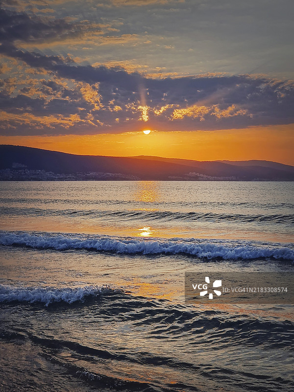 保加利亚黑海海岸的生机勃勃的日出。阳光明媚的海滩度假胜地，黎明时分，太阳在地平线的山丘上升起，泡沫波浪拍打着沙滩。图片素材